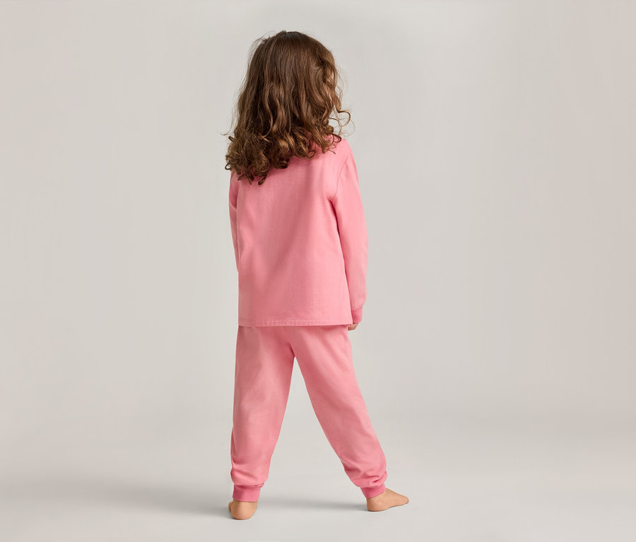 Un enfant aux cheveux bouclés se tient dos à l'objectif sur un fond gris clair, vêtu d'un ensemble de pyjama rose.