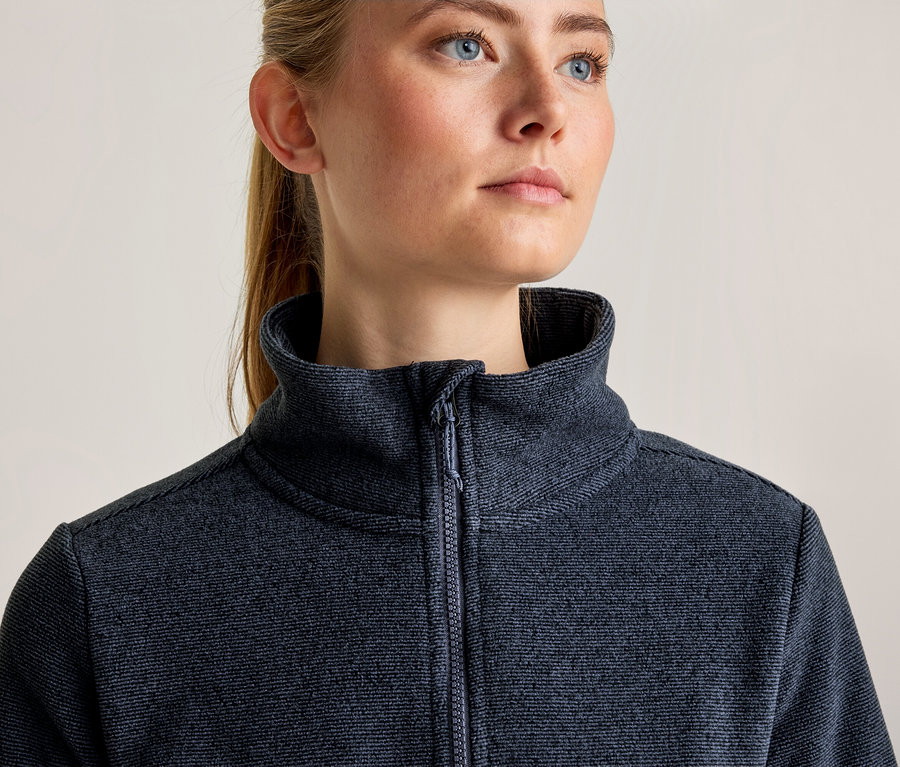 Gros plan d'une femme aux cheveux blonds et aux yeux bleus, regardant vers la droite, portant une veste zippée texturée bleu foncé avec un col montant.