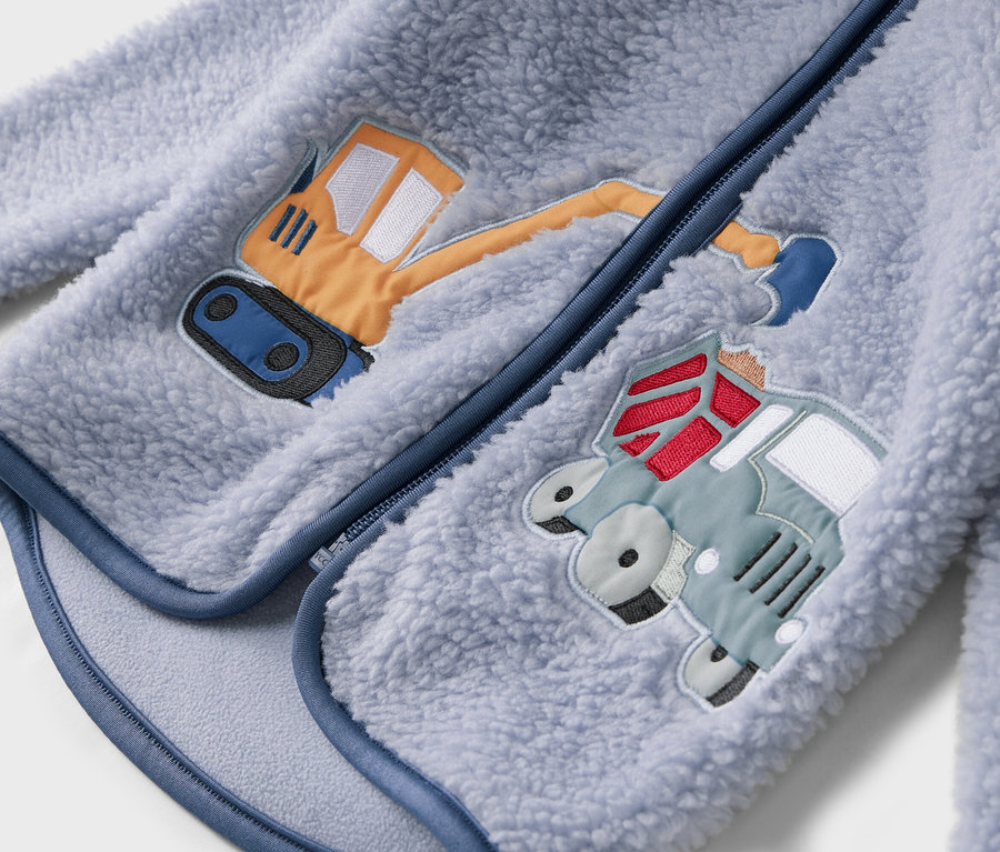 Gros plan sur une veste polaire bleu clair avec fermeture éclair et motifs cousus représentant une excavatrice et un camion.