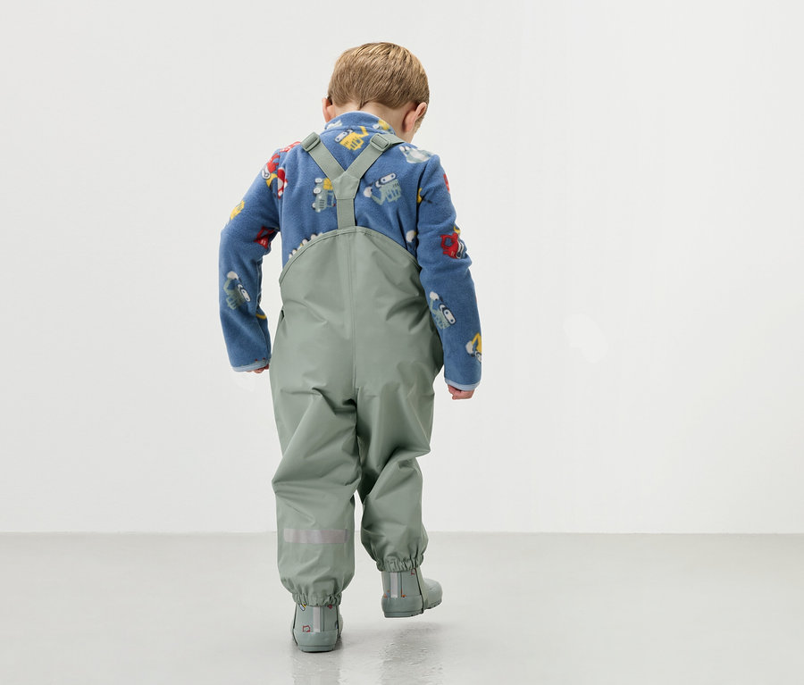 Enfant vu de dos, portant une veste polaire bleue à motifs de véhicules, un pantalon de pluie vert et des bottes en caoutchouc.