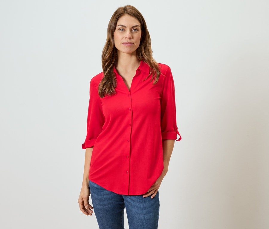 Une femme aux cheveux bruns porte un chemisier rouge et un jean bleu.
