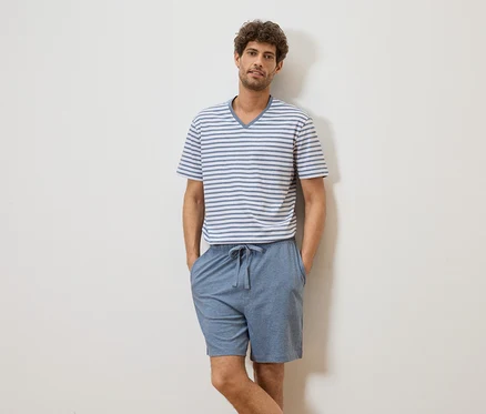 Un homme aux cheveux bouclés portant un t-shirt rayé et un short bleu se tient devant un mur blanc.