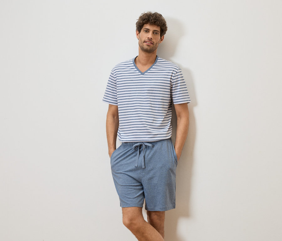 Un homme aux cheveux bouclés portant un t-shirt rayé et un short bleu se tient devant un mur blanc.