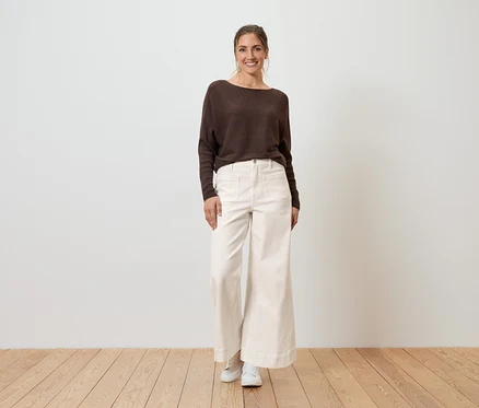 Une femme se tient devant un mur, portant un pull marron, un pantalon ample et clair et des chaussures blanches.
