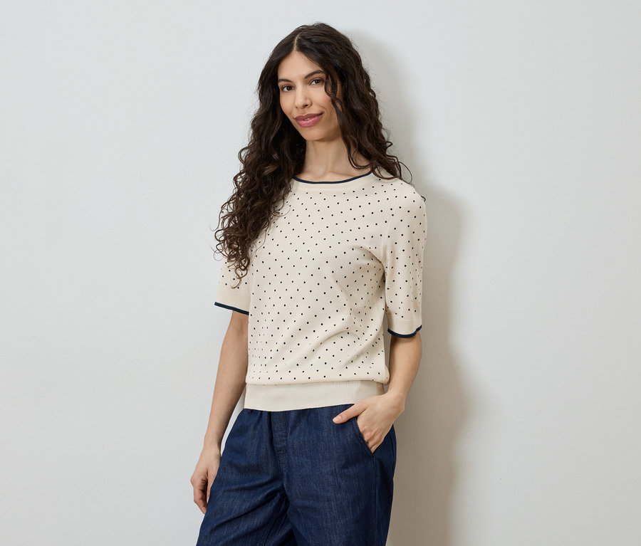 Une femme aux cheveux bouclés porte un haut clair à manches courtes à pois noirs et un pantalon bleu devant un mur blanc.