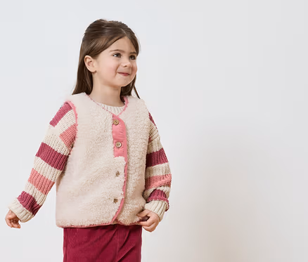 Fillette souriante portant un pull rayé et un gilet beige avec des touches de rose sur fond blanc.