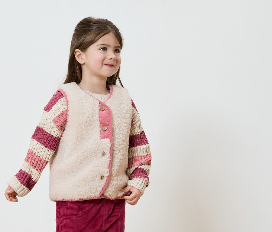 Fillette souriante portant un pull rayé et un gilet beige avec des touches de rose sur fond blanc.