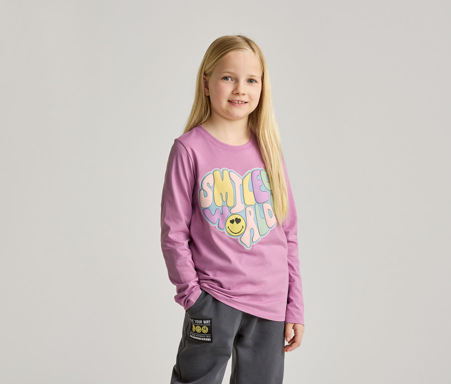 Une fillette aux longs cheveux blonds porte un t-shirt violet à manches longues avec un imprimé cœur coloré et un pantalon de jogging gris.