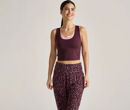 Une femme souriante portant un débardeur bordeaux sur un haut rose et un pantalon à imprimé léopard sur un fond clair.