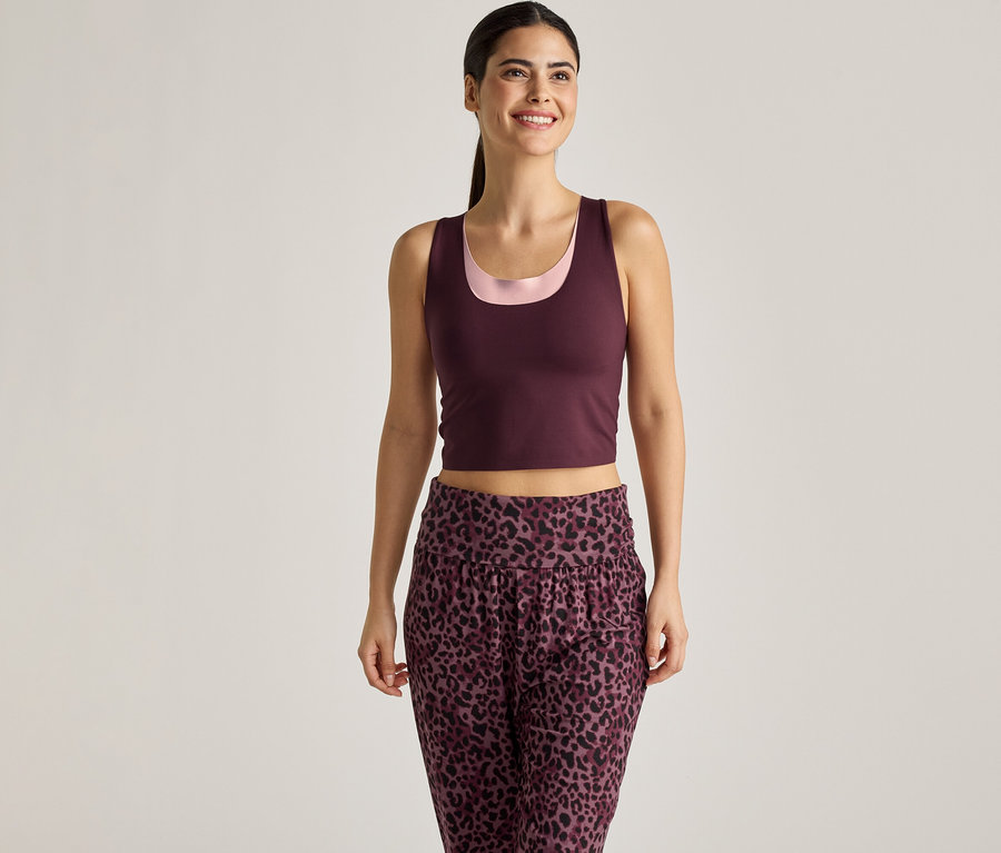Une femme souriante portant un débardeur bordeaux sur un haut rose et un pantalon à imprimé léopard sur un fond clair.
