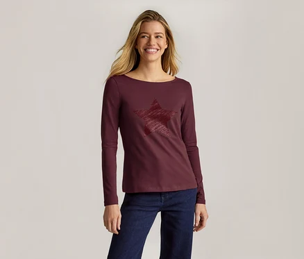 Femme souriante aux longs cheveux blonds portant un haut bordeaux à manches longues avec un motif étoile texturé et un jean bleu foncé.