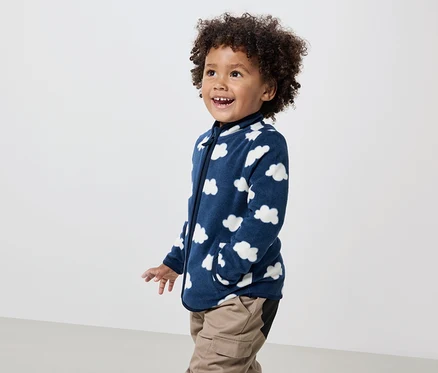 Un garçon souriant aux cheveux bouclés porte une veste polaire bleu marine à motifs de nuages et un pantalon kaki.