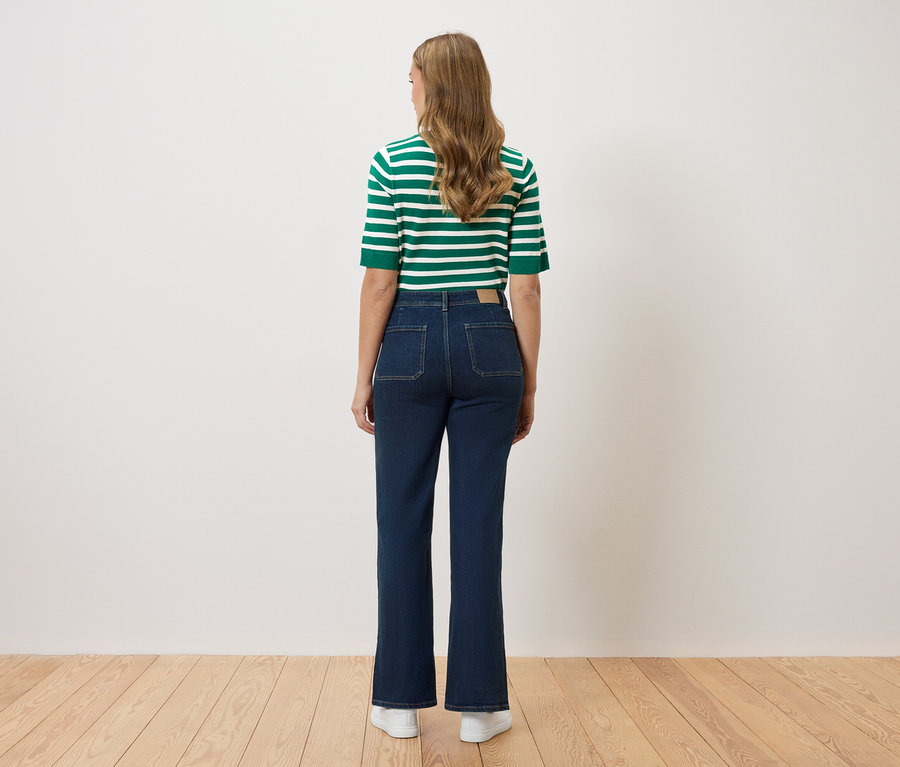 Vue de dos d'une femme aux longs cheveux ondulés portant un haut rayé vert et blanc, un jean bleu marine évasé et des baskets blanches.