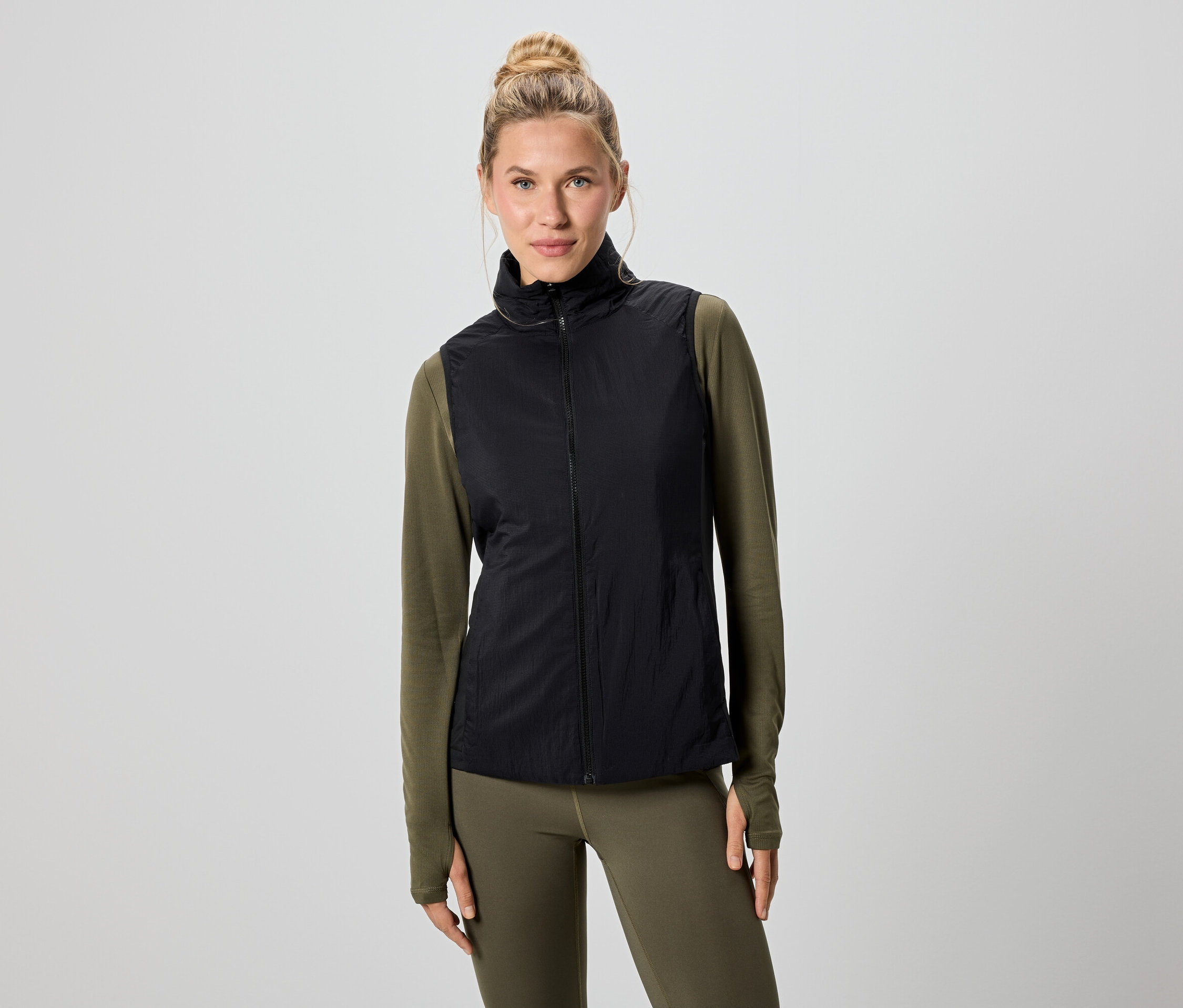 Une femme blonde porte un gilet noir sur un t-shirt vert à manches longues et un legging.