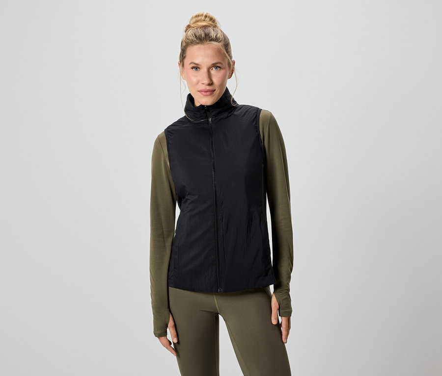 Une femme blonde porte un gilet noir sur un t-shirt vert à manches longues et un legging.