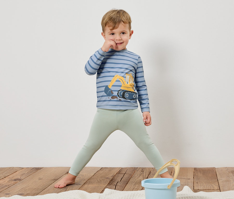 Un garçon se tient pieds nus sur un plancher en bois, portant un t-shirt rayé à manches longues avec une pelle mécanique et un legging vert clair. Un seau bleu est posé devant lui.