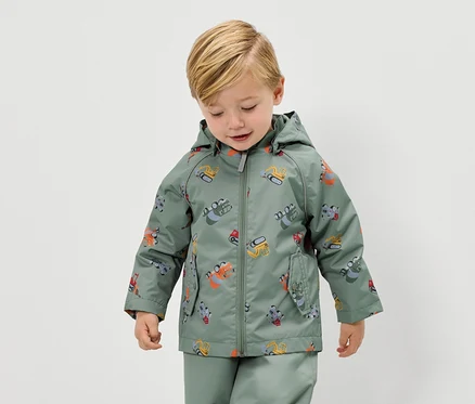 Un garçon blond porte un imperméable vert à motif d'excavatrice et un pantalon de pluie assorti.