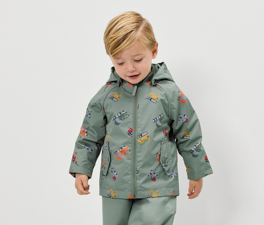 Un garçon blond porte un imperméable vert à motif d'excavatrice et un pantalon de pluie assorti.