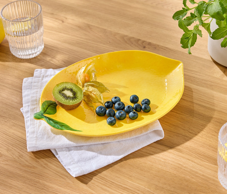 Une assiette jaune avec des myrtilles, un kiwi et des physalis est posée sur un linge blanc. À côté, il y a un verre d'eau avec du citron et un citron.