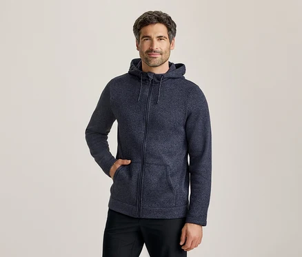 Un homme portant un sweat à capuche zippé bleu foncé, une main dans la poche, debout devant un fond clair.