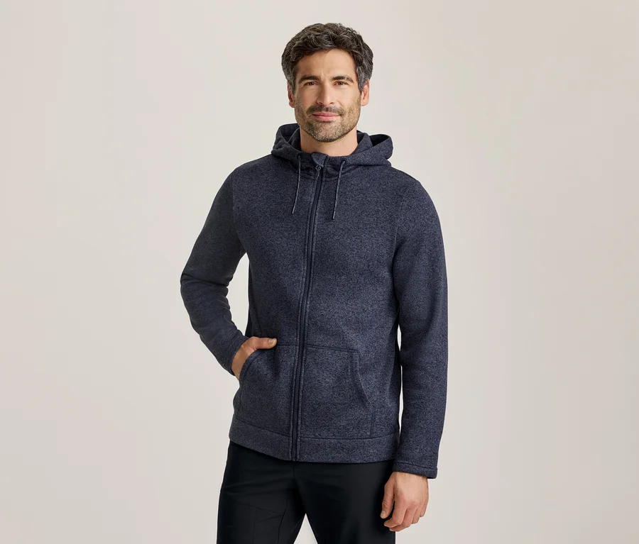 Un homme portant un sweat à capuche zippé bleu foncé, une main dans la poche, debout devant un fond clair.