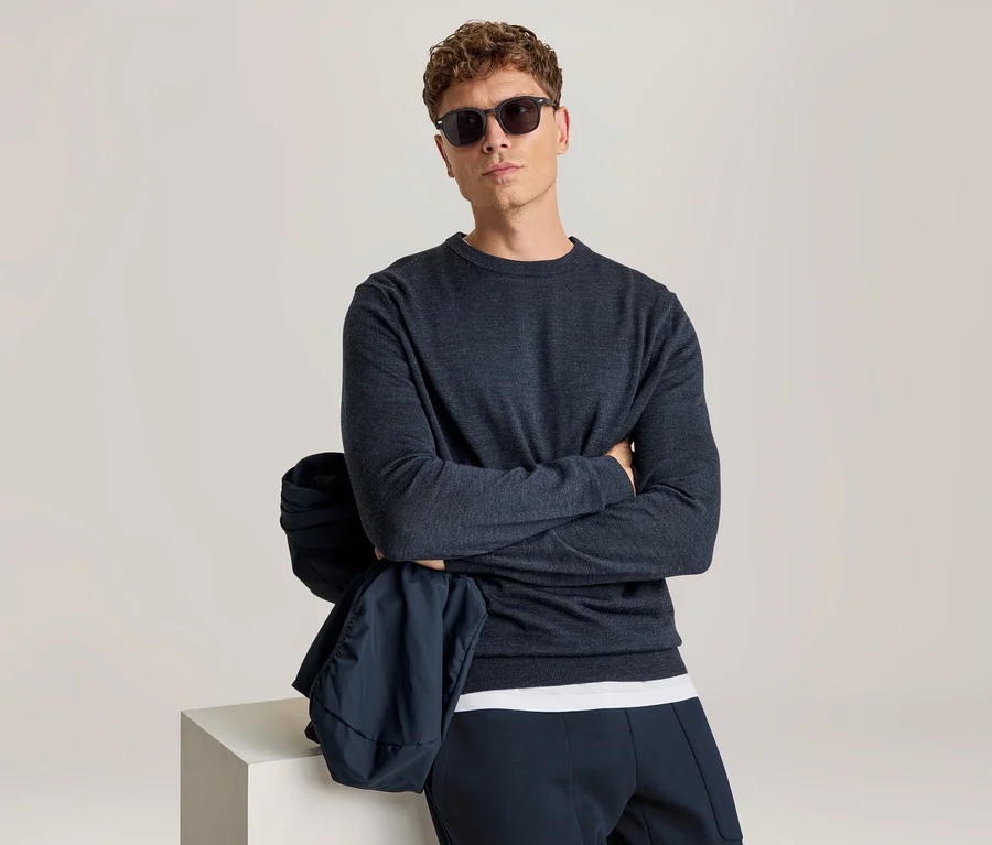 Homme aux cheveux bruns bouclés et lunettes de soleil, regardant directement l'appareil photo. Il porte un haut à manches longues bleu foncé, un t-shirt blanc en dessous et un pantalon bleu foncé. Ses bras sont croisés et une veste bleu foncé est drapée sur son bras gauche, reposant sur un cube blanc.