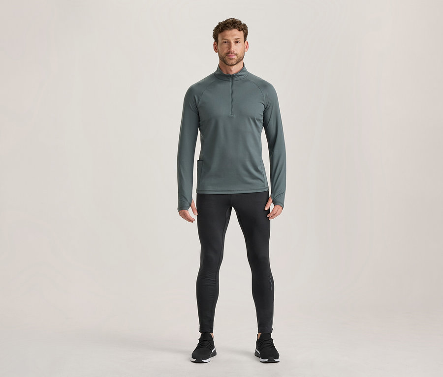 Homme debout portant un haut de sport gris foncé à manches longues avec fermeture éclair et passe-pouces, un legging de sport noir et des chaussures de sport noires.