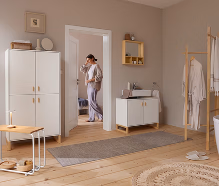 Une femme entre dans une salle de bain lumineuse avec un parquet en bois. À droite, une échelle à vêtements en bois tient un peignoir et des serviettes à côté d'une baignoire blanche. Au-dessus d'un meuble-lavabo blanc avec un lavabo, un miroir avec étagères est fixé au mur. À gauche, une grande armoire blanche se trouve à côté d'une petite table avec une lampe.