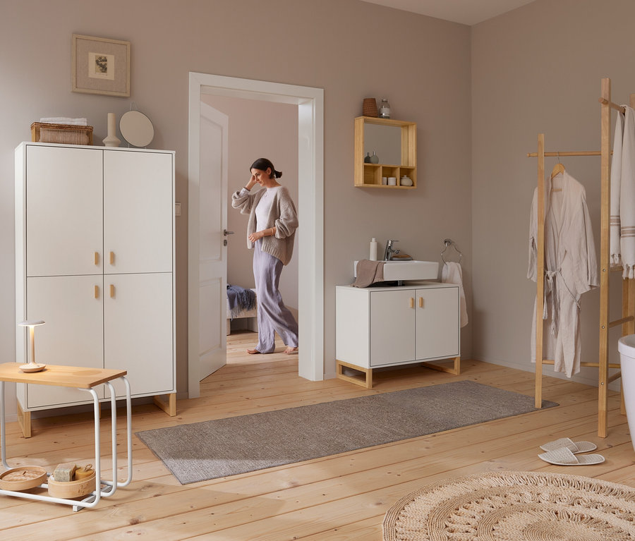 Une femme entre dans une salle de bain lumineuse avec un parquet en bois. À droite, une échelle à vêtements en bois tient un peignoir et des serviettes à côté d'une baignoire blanche. Au-dessus d'un meuble-lavabo blanc avec un lavabo, un miroir avec étagères est fixé au mur. À gauche, une grande armoire blanche se trouve à côté d'une petite table avec une lampe.