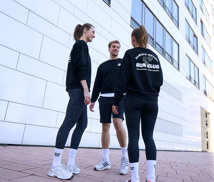 Trois personnes, deux femmes et un homme, se tiennent debout, souriantes et discutant. Tous portent des sweatshirts noirs et des chaussettes de tennis Unisex Coffee Crew blanches. L'homme porte un short de sport noir. Une femme porte des leggings de sport « ActivePower » gris-noir, l'autre des leggings de sport « ActivePower » noirs.