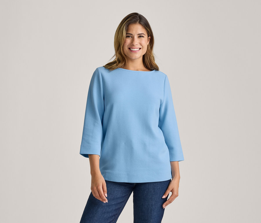 Femme souriante portant un haut bleu clair à col bateau et manches trois-quarts avec un jean bleu foncé, debout devant un fond clair.