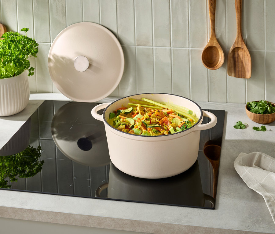 Une cocotte beige remplie de légumes colorés comme du brocoli, des carottes et du céleri, est posée sur une plaque à induction noire. À côté se trouvent un couvercle assorti, du persil frais, des ustensiles en bois et de la coriandre sur le comptoir de la cuisine.