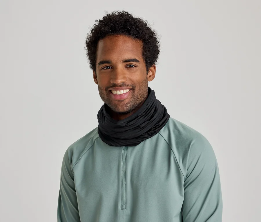 Un homme souriant aux cheveux bouclés porte un cache-cou noir et un t-shirt à manches longues vert clair.