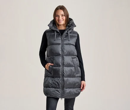 Femme souriante portant une longue doudoune sans manches gris foncé à capuche sur un haut noir à manches longues et un pantalon foncé, une main dans une poche de la veste.