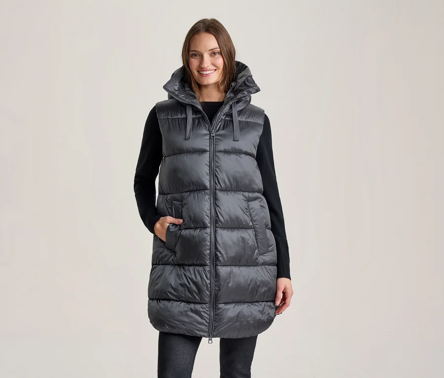 Femme souriante portant une longue doudoune sans manches gris foncé à capuche sur un haut noir à manches longues et un pantalon foncé, une main dans une poche de la veste.