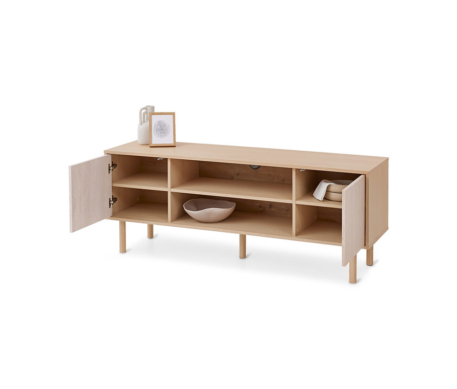 Un buffet bas en bois clair avec des étagères ouvertes et deux portes légèrement entrouvertes. Sur le dessus, un vase blanc et un cadre photo. À l'intérieur, un bol en bois et des plats blancs avec un tissu.