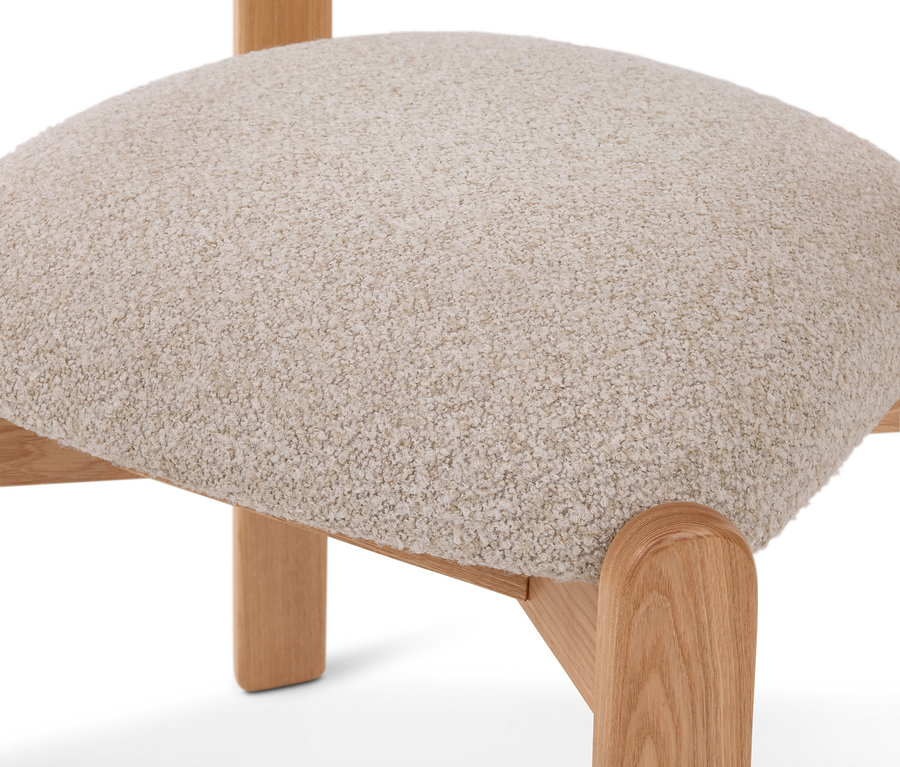 Gros plan d'une assise de chaise recouverte de tissu texturé beige clair, soutenue par des pieds en bois clair aux bords arrondis.