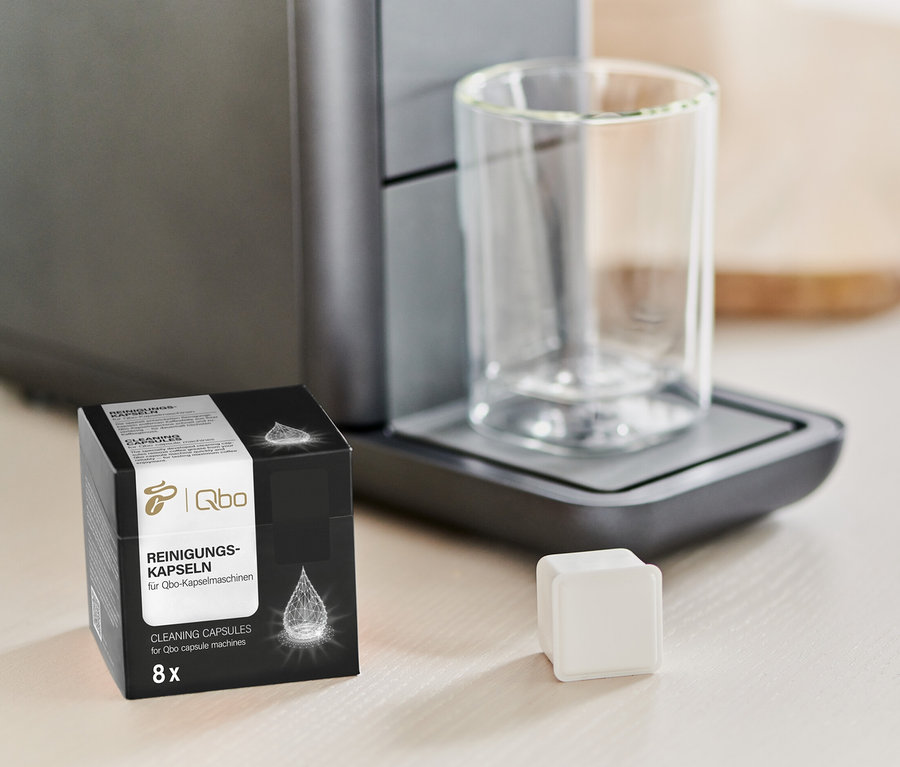 Un paquet de capsules de nettoyage Qbo est placé à côté d'une machine à café et d'un verre.