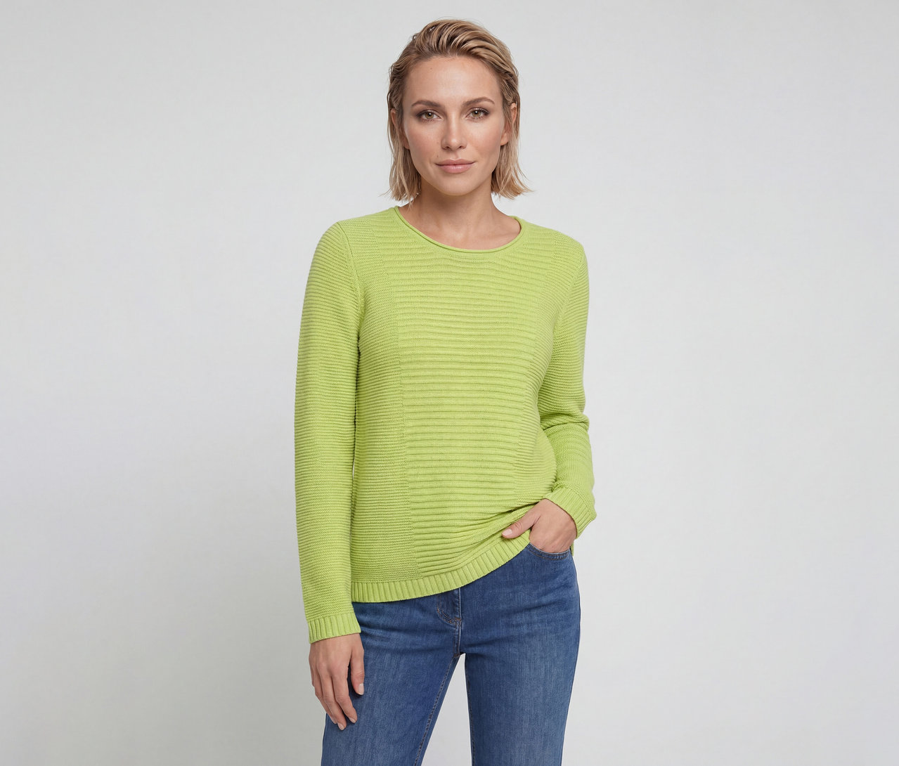 Femme aux cheveux blonds courts portant un pull côtelé vert citron et un jean bleu, une main dans la poche, regardant l'appareil photo.