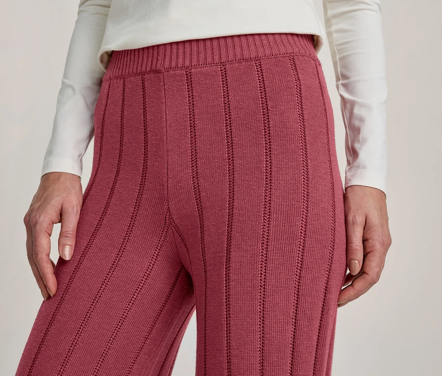 Gros plan d'une personne portant un haut blanc à manches longues et un pantalon en maille côtelée couleur baie. Leurs mains sont posées sur les côtés.