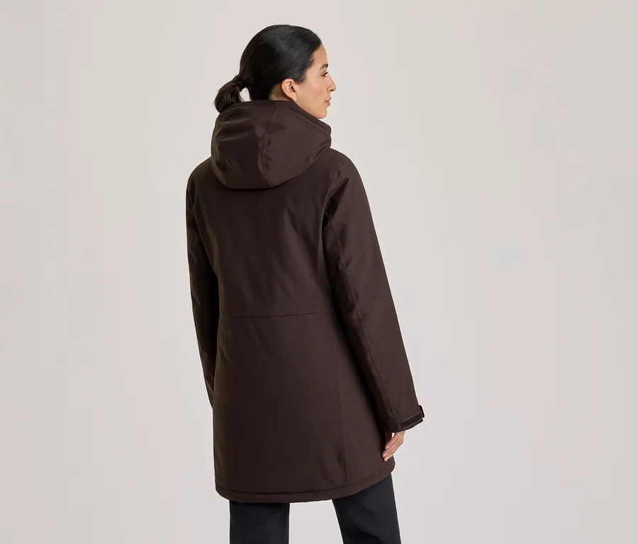 Femme vue de dos, légèrement tournée vers la droite, portant une parka à capuche marron foncé et un pantalon sombre. Ses cheveux sont attachés en queue de cheval.