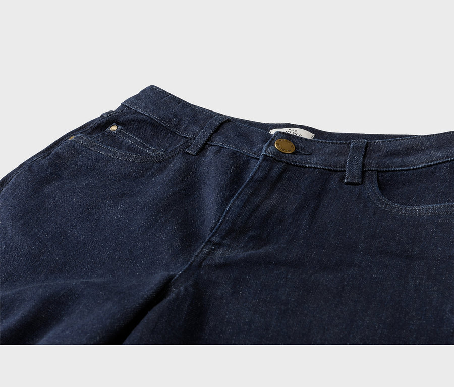 Gros plan sur un jean en denim bleu foncé, montrant la ceinture, les passants de ceinture, les poches avant, une petite poche à monnaie avec un rivet en laiton et une fermeture boutonnée en laiton.