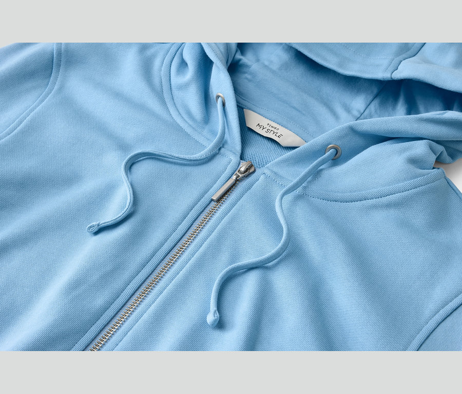 Gros plan d'un sweat à capuche zippé bleu clair avec une fermeture éclair argentée et des cordons de serrage assortis. Une étiquette blanche avec « TCHIBO MY STYLE » est visible.