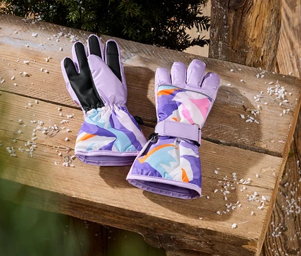 Deux gants violets avec un motif abstrait coloré sont posés sur une table en bois avec de la neige artificielle et des branches de sapin.