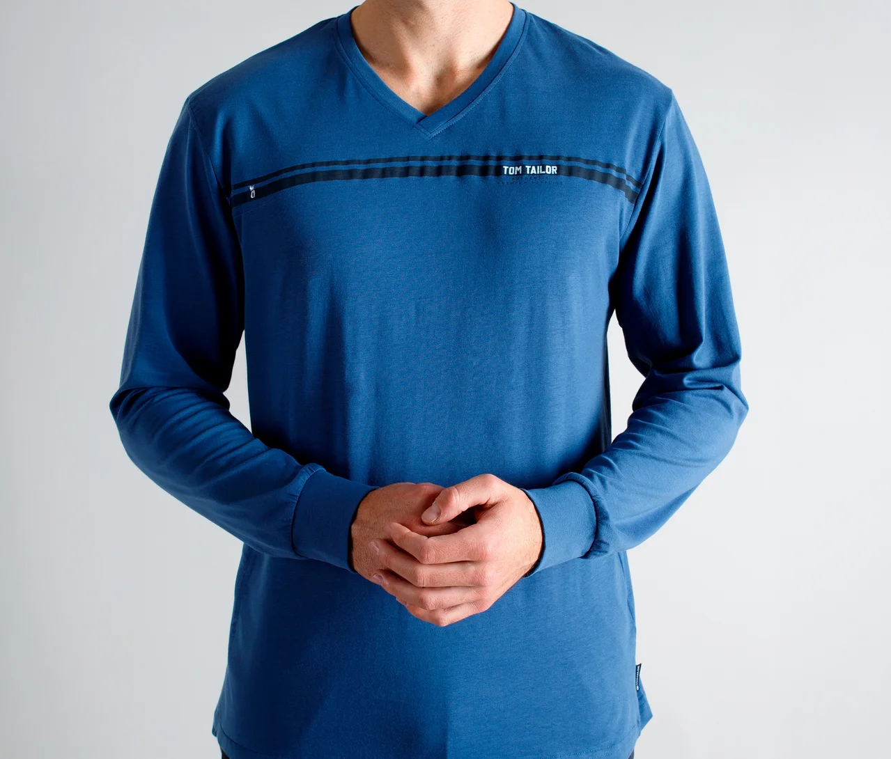 Homme portant un t-shirt bleu à manches longues et col en V, avec des rayures noires et le texte « TOM TAILOR » sur la poitrine, les mains jointes.