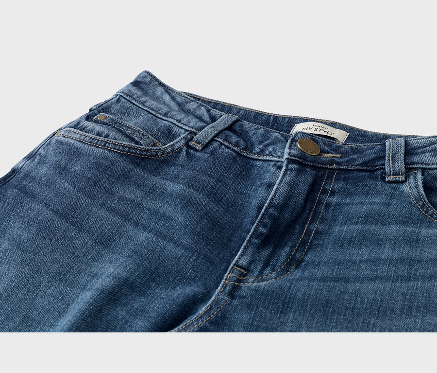 Gros plan sur un jean en denim bleu montrant la taille, le bouton avant, les passants de ceinture et les poches. Une étiquette blanche avec « TCHIBO MY STYLE » est visible près du bouton en laiton.
