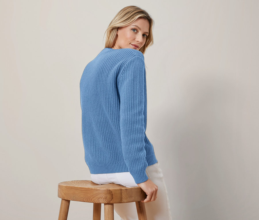 Une femme aux cheveux blonds porte un cardigan bleu en grosse maille et un pantalon blanc, assise sur un tabouret en bois, regardant par-dessus son épaule gauche.