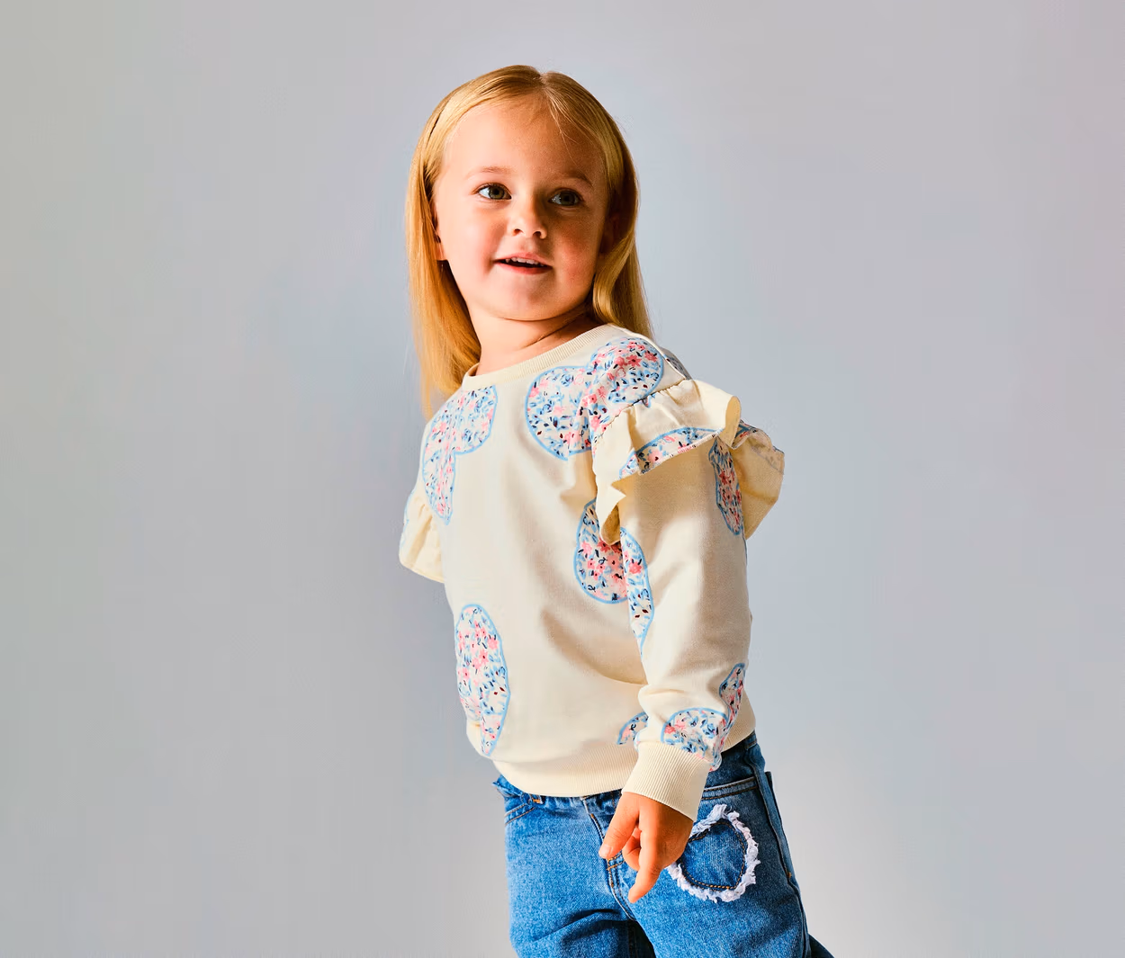 Jeune fille aux cheveux blonds portant un pull NAME IT Kinder-Sweater crème avec des motifs de cœurs floraux et des volants aux épaules, un jean bleu, regardant vers la droite.
