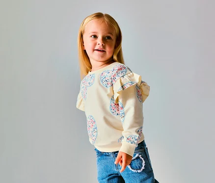 Jeune fille aux cheveux blonds portant un pull NAME IT Kinder-Sweater crème avec des motifs de cœurs floraux et des volants aux épaules, un jean bleu, regardant vers la droite.