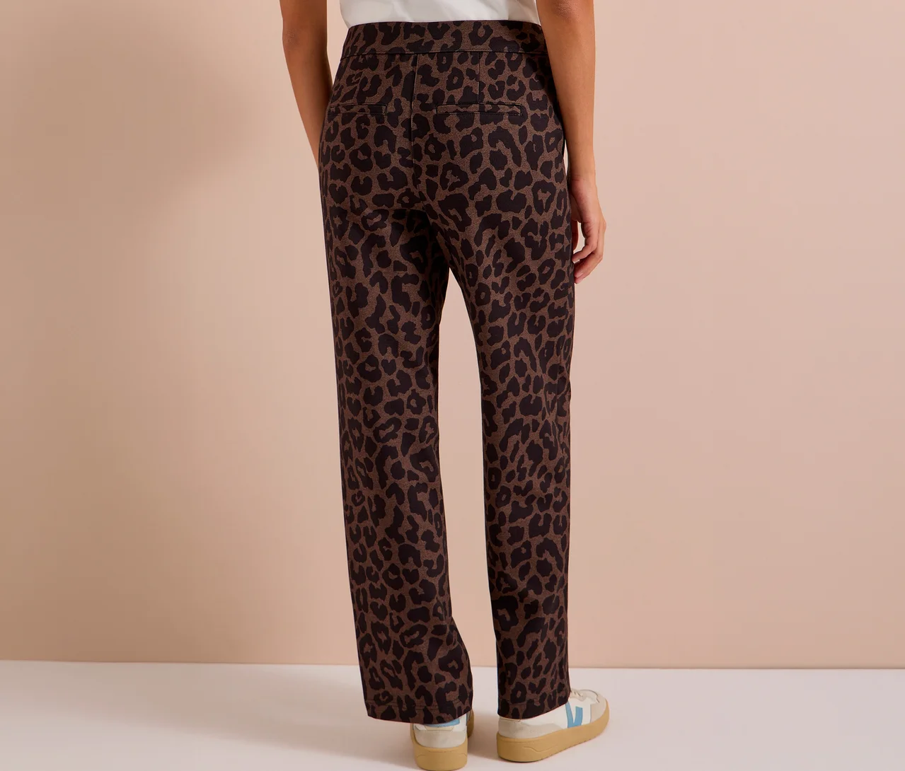 Personne vue de dos portant un pantalon en tissu CECIL marron et noir à imprimé léopard sur toute la surface, avec des baskets blanches et beiges.